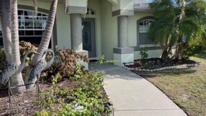 A two-story house with a light green exterior, a covered front porch, and a well-maintained garden with palm trees and shrubs. - Rotonda Heights Pool Home (Rotonda West)