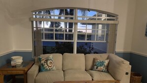 A living room with a beige sofa, a glass-top coffee table, a side table, and a large window with a view of palm trees. - Rotonda Heights Pool Home (Rotonda West)