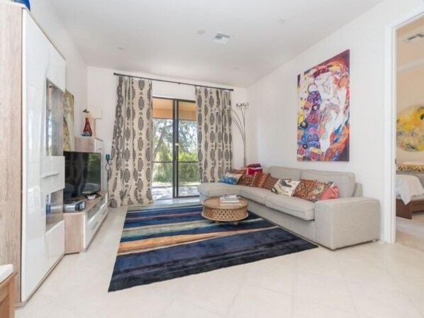 Un salón moderno con sofá, mesa de centro y televisor. Hay un ventanal con cortinas y un cuadro de colores en la pared.