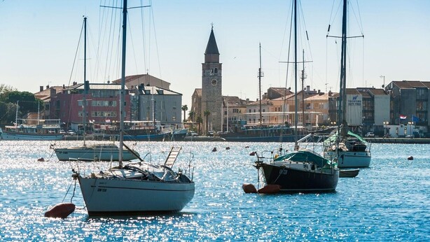A marina with several sailboats docked, a prominent tower in the background, and buildings along the waterfront. - Sea View Studio Superior (Umag)
