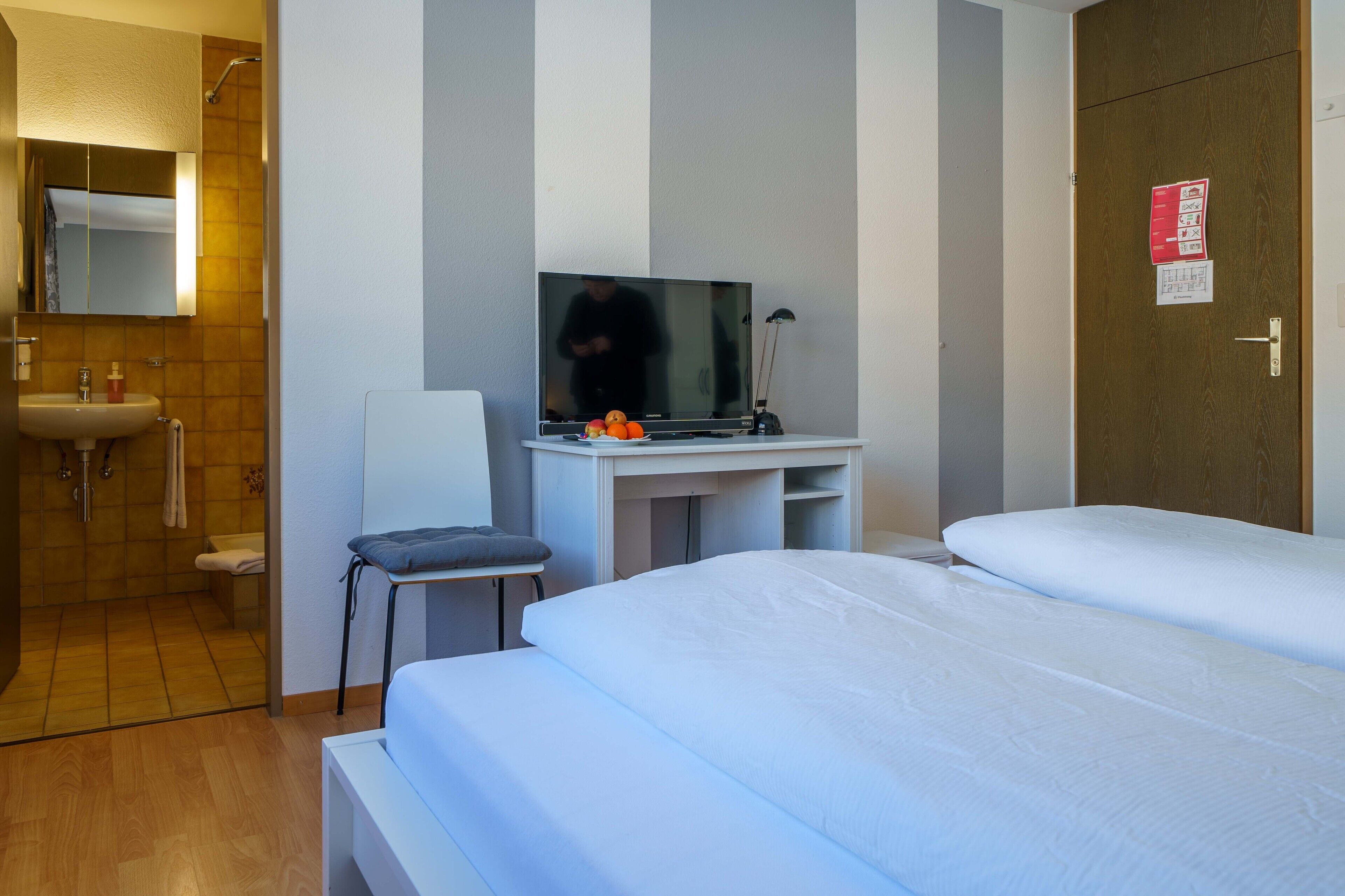 A hotel room with a bed, a desk with a television, a chair, a sink, and a shower.