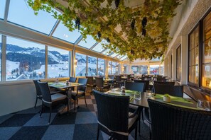 A restaurant with large windows, wicker furniture, and a view of snow-covered mountains. - Hotel Toggenburg (Wildhaus-Alt St. Johann)