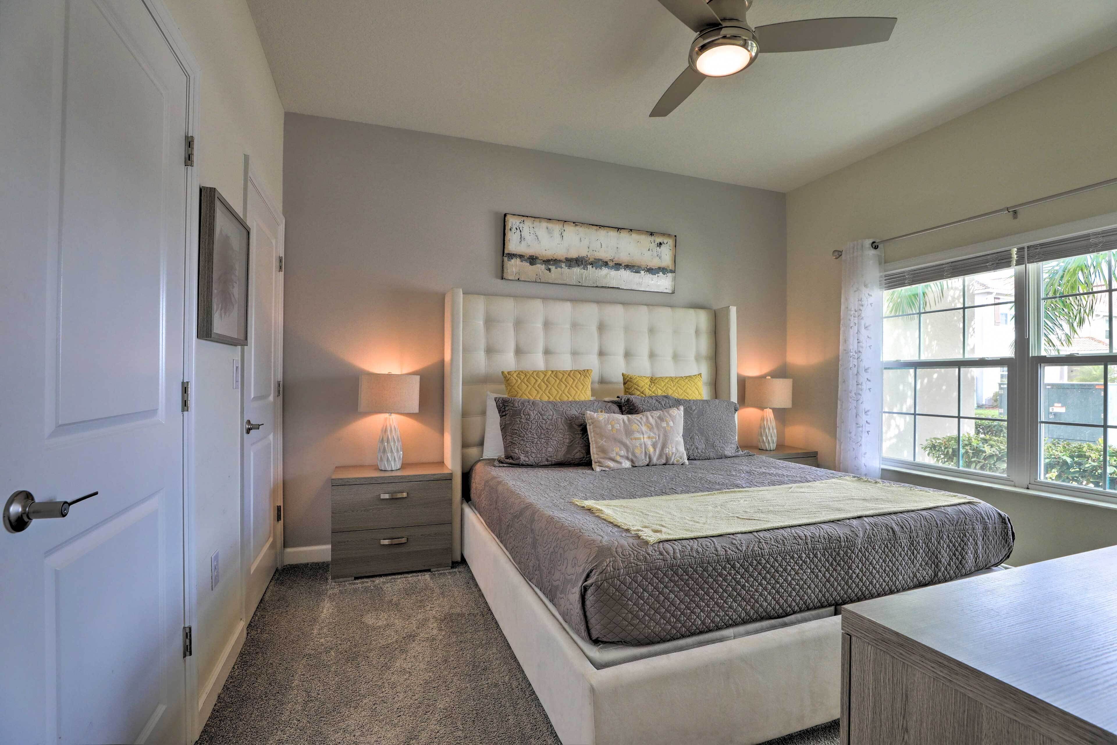 A bedroom with a tufted headboard, a large window with a view, a ceiling fan, and a bedside table with a lamp.