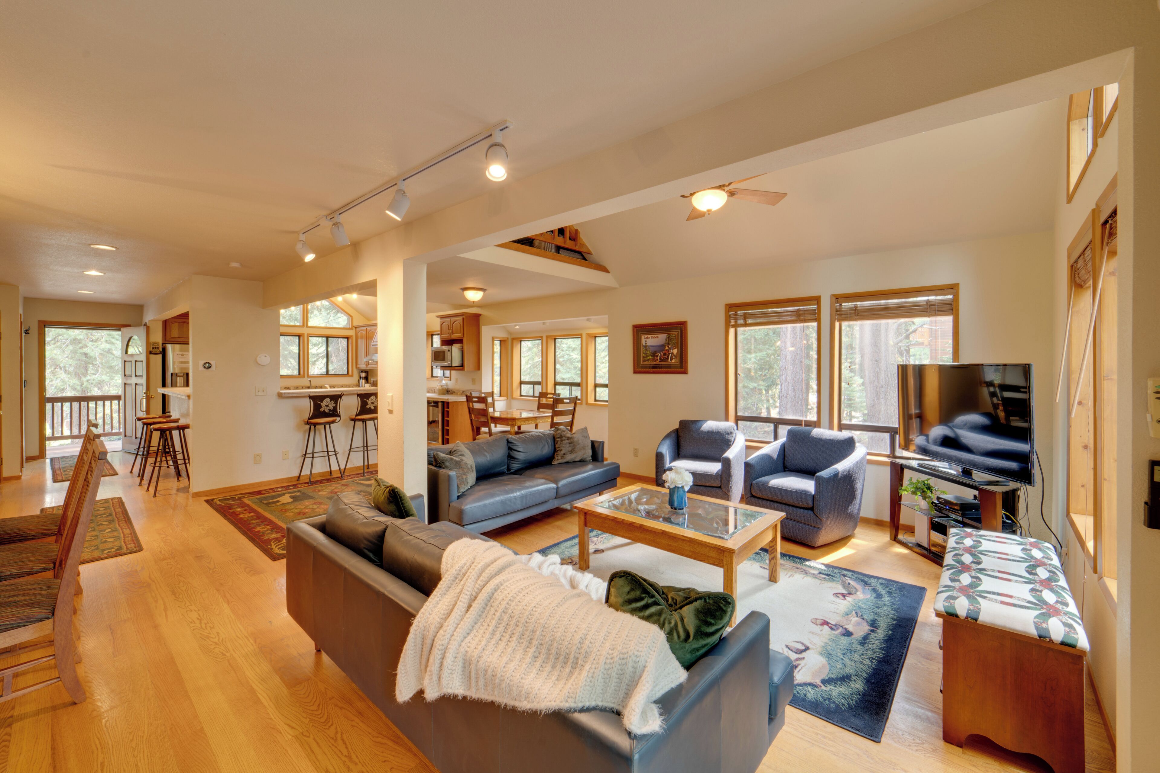 A spacious living room with a sofa, chairs, a coffee table, and a television. There is a dining area with a table and chairs, and a kitchen in the background.