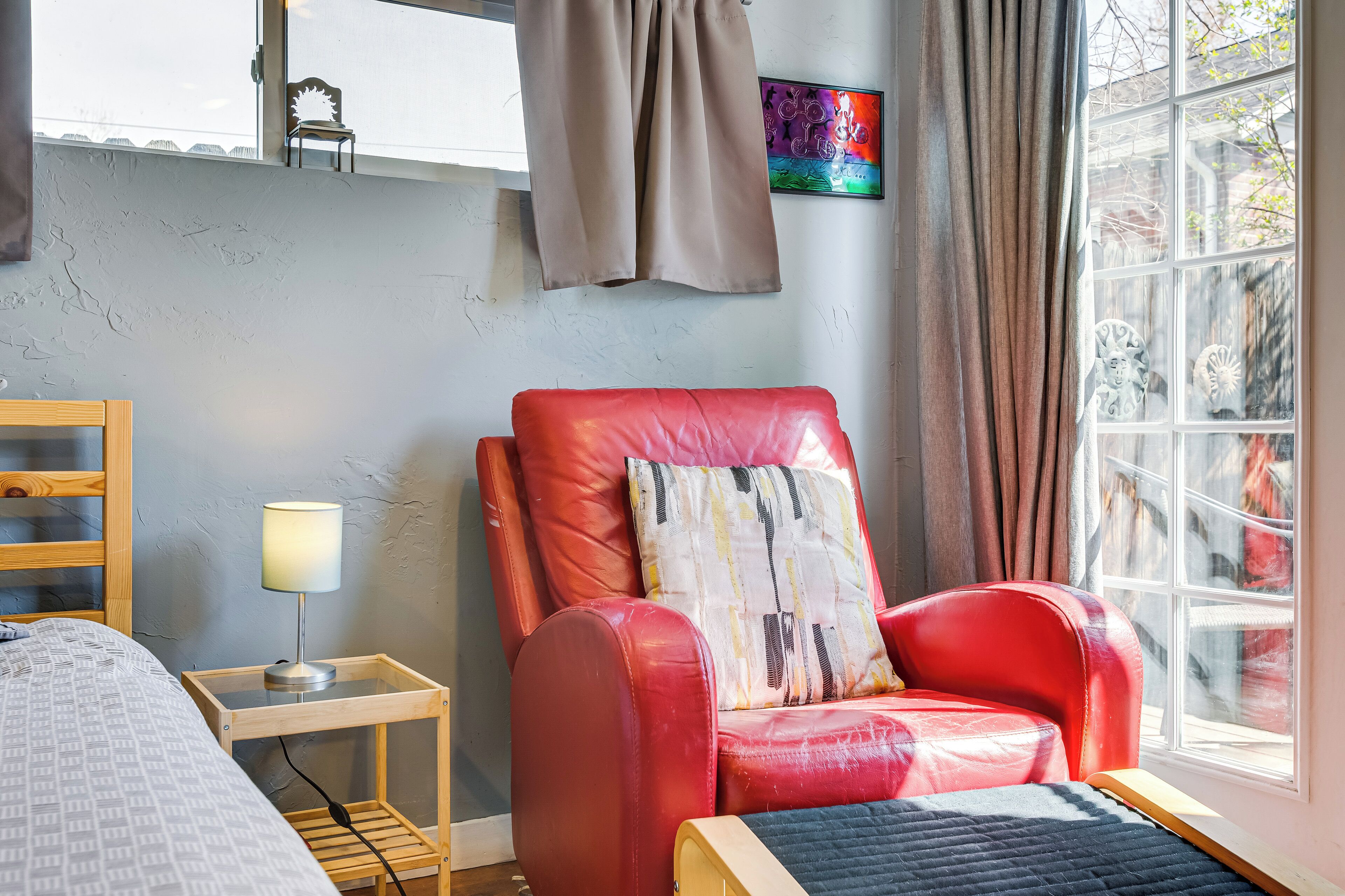 Ein Zimmer mit einem roten Ledersessel, einem Holztischchen mit einer Lampe, einem Bett mit einem gemusterten Überwurf und einem Fenster mit Vorhängen.