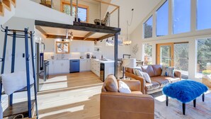 Een ruime woonkamer met een hoge plafond, een keuken met witte kastjes en een groot raam met uitzicht op een tuin.