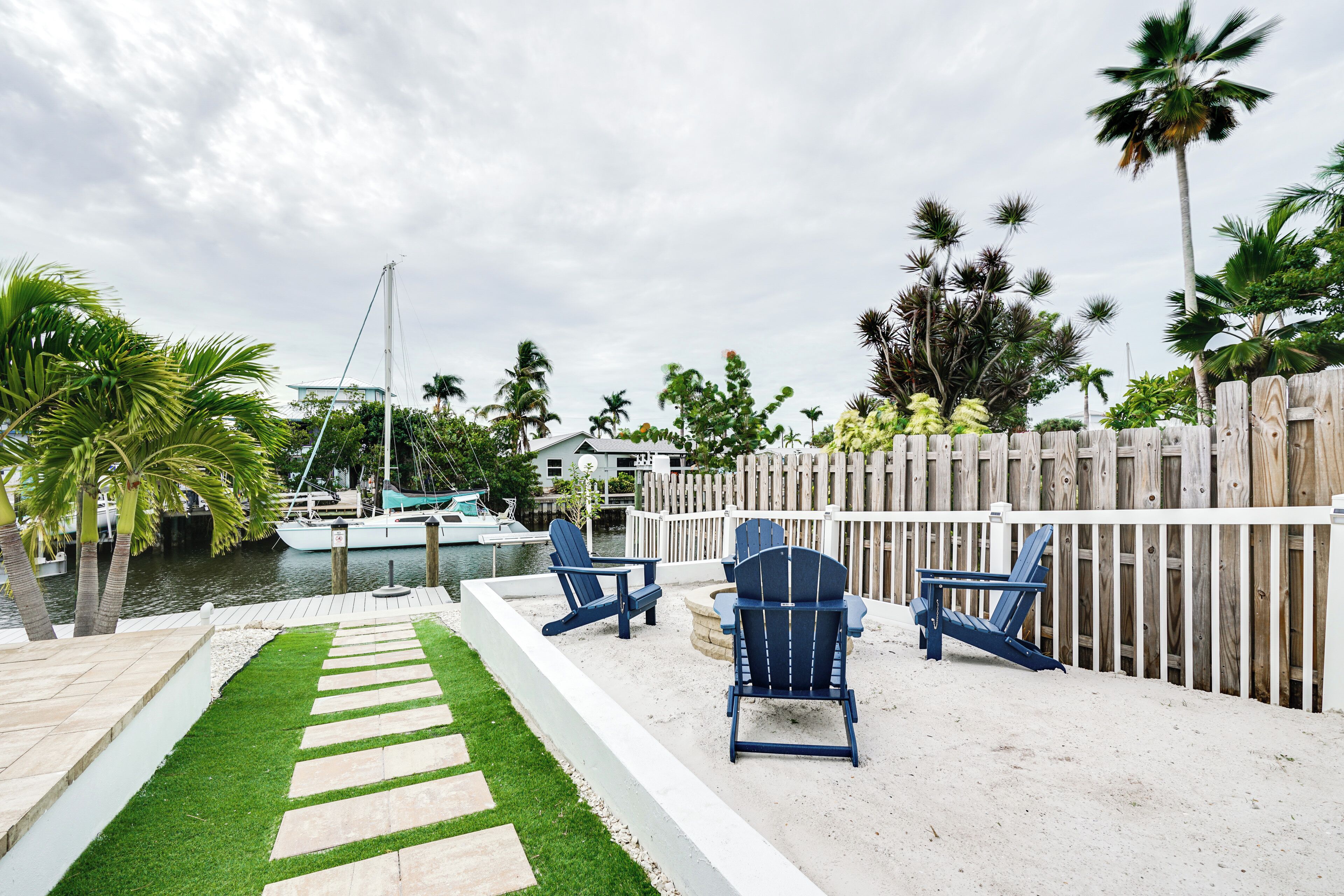 A patio area with a wooden fence, two blue Adirondack chairs, a stone pathway, and a view of a marina with boats.