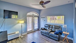 A living room with a ceiling fan, a flat-screen TV, a leather sofa, a lamp, and a window with blinds. - Naples Studio w/ Resort Amenities - Near Marina! (Naples)