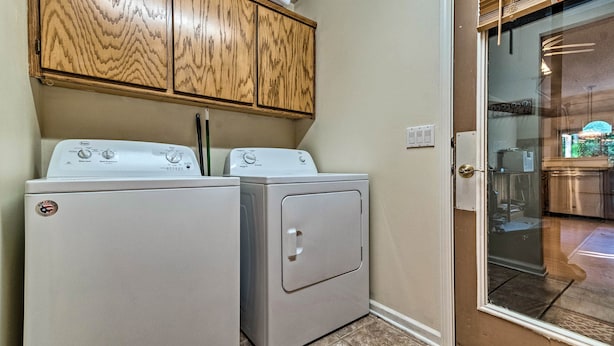 A laundry area with two washing machines and a dryer, wooden cabinets above, and a glass door leading to a kitchen area. - Hot Springs Village Home, Walk to Lake & Golf (Hot Springs Village)