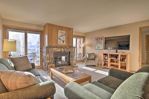 A cozy living room with a stone fireplace, green upholstered sofas, a wooden coffee table, and a television on a wooden stand. - Scenic Buffalo Village Condo w/ View & Pool Access (Silverthorne)