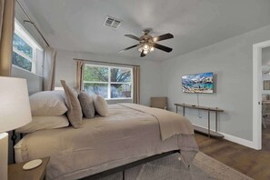 A bedroom with a bed, a ceiling fan, a window with curtains, a bedside table, and a painting on the wall. - Weitz Retreat with Private Hot Tub (Fredericksburg)
