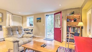 Un salon comprenant un fauteuil inclinable en cuir, une table basse en bois, une bibliothèque et une porte rouge donnant sur l’extérieur.