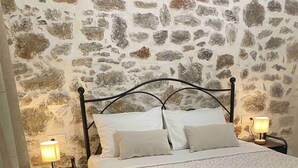 Un dormitorio con pared de piedra, una cama con ropa de cama blanca, dos mesitas de noche con lámparas y suelo de madera.