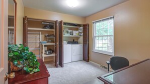 A laundry room with a washing machine, dryer, shelves with various items, and a window with blinds. - Family-friendly West Chester Twp Home w/ Pool! (West Chester)