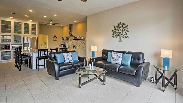 A modern living room with a glass coffee table, two sofas with blue cushions, a dining area with chairs, and a kitchen in the background. - Cape Coral House w/ Private Dock, Pool & Bar! (Cape Coral)