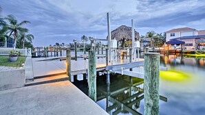 A waterfront dock area with a thatched-roof structure, wooden posts, and a calm water body. - Cape Coral House w/ Private Dock, Pool & Bar! (Cape Coral)