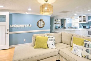 A living room with a beige sofa, a hanging light fixture, and a kitchen in the background. - Walk to Ocean: Home w/ Fire Pit in Atlantic Beach (North Myrtle Beach)