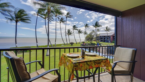 A balcony with a table set for two, overlooking a grassy area with palm trees and a view of the sea. - Molokai Shores Resort Condo w/ Pool & Views! (Kaunakakai)