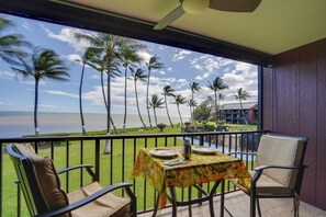 A balcony with a table set for two, overlooking a grassy area with palm trees and a view of the sea. - Molokai Shores Resort Condo w/ Pool & Views! (Kaunakakai)