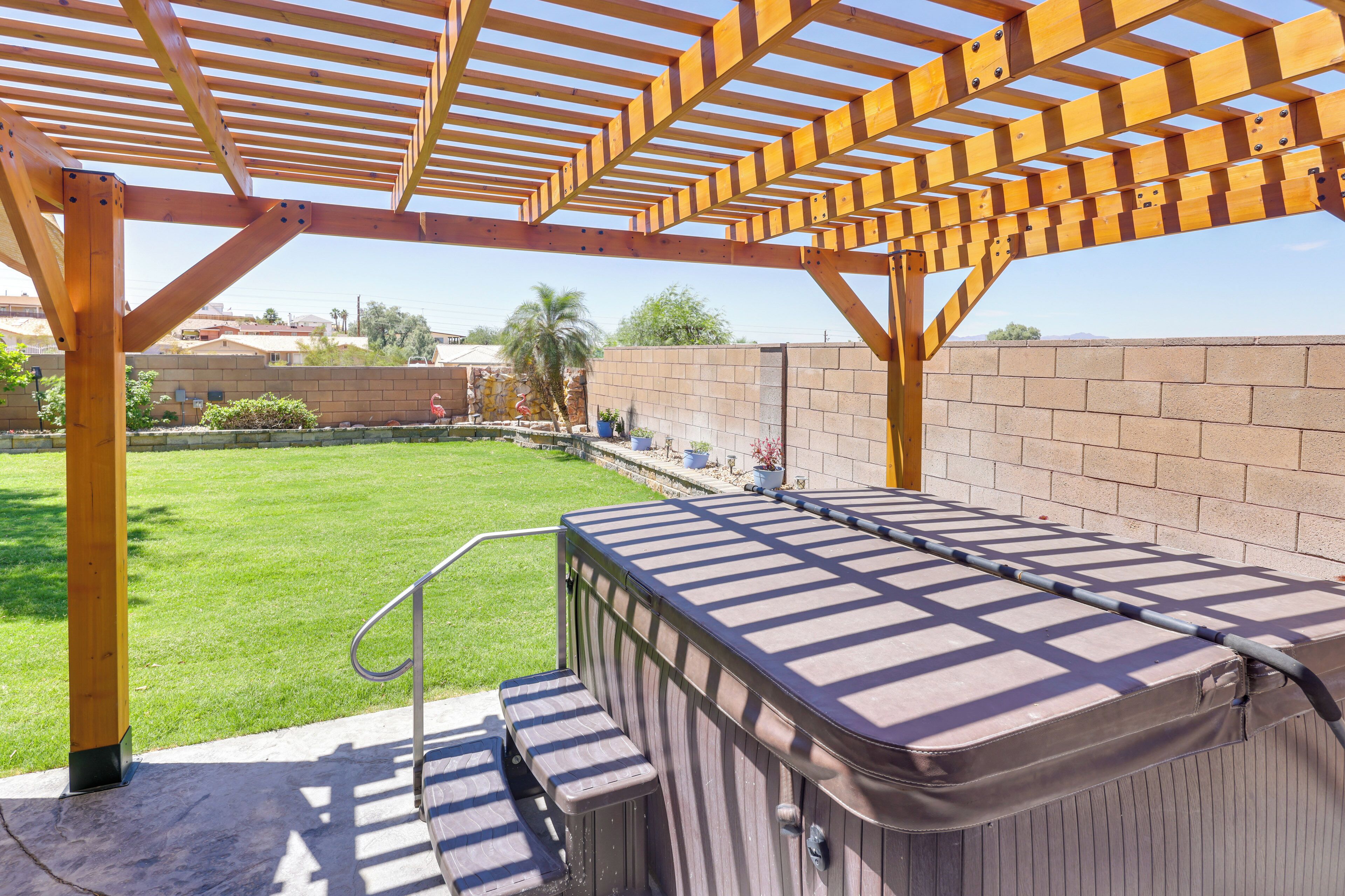 A hot tub under a wooden pergola in a backyard with a brick wall and a grassy lawn.