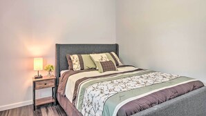 Un lit bien fait, recouvert d’une couvre-lit à motifs floraux et agrémenté de plusieurs coussins, une table de chevet avec une lampe et un vase de fleurs, et une tête de lit sombre.