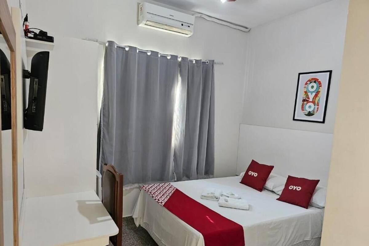 A hotel room with a bed, a desk, a chair, a window with curtains, an air conditioning unit, and a framed picture on the wall.
