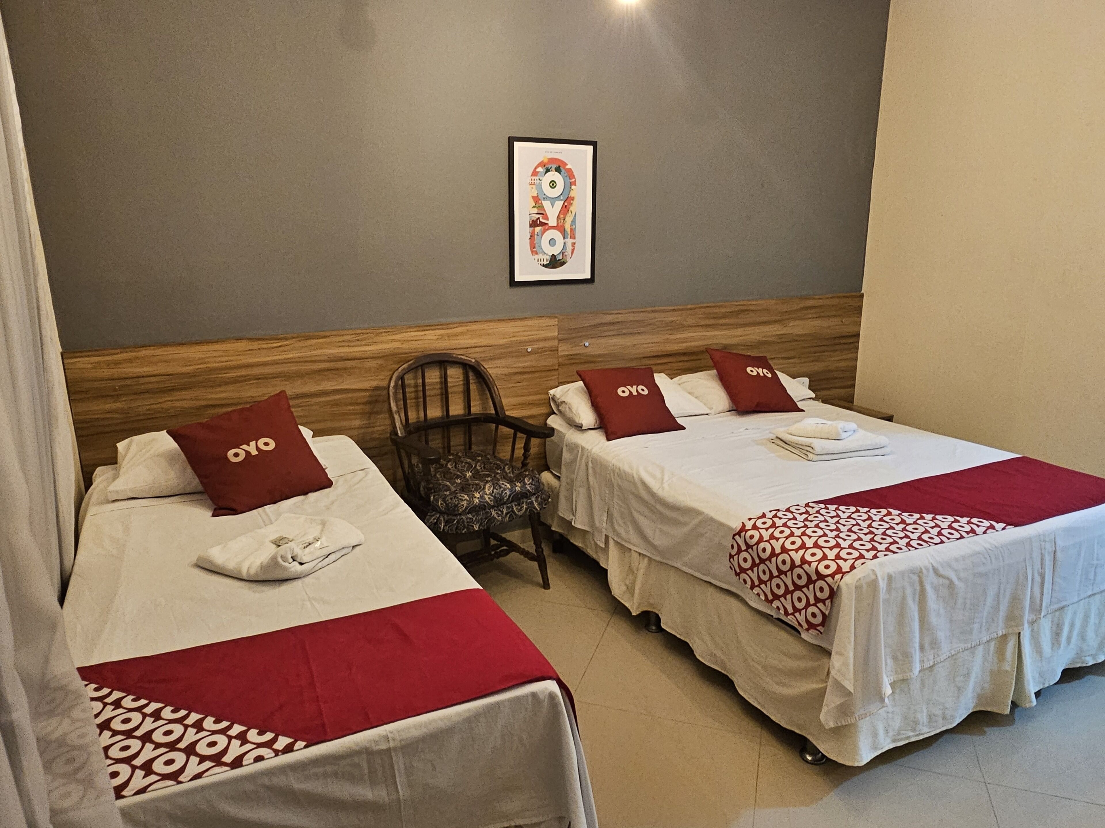 A hotel room with two beds, each with a red and white bedspread and matching pillows. There is a wooden headboard, a chair with a patterned cushion, and a framed picture on the wall.