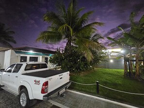 Una camioneta pickup blanca estacionada frente a un edificio con techo verde, rodeado de vegetación y una palmera.