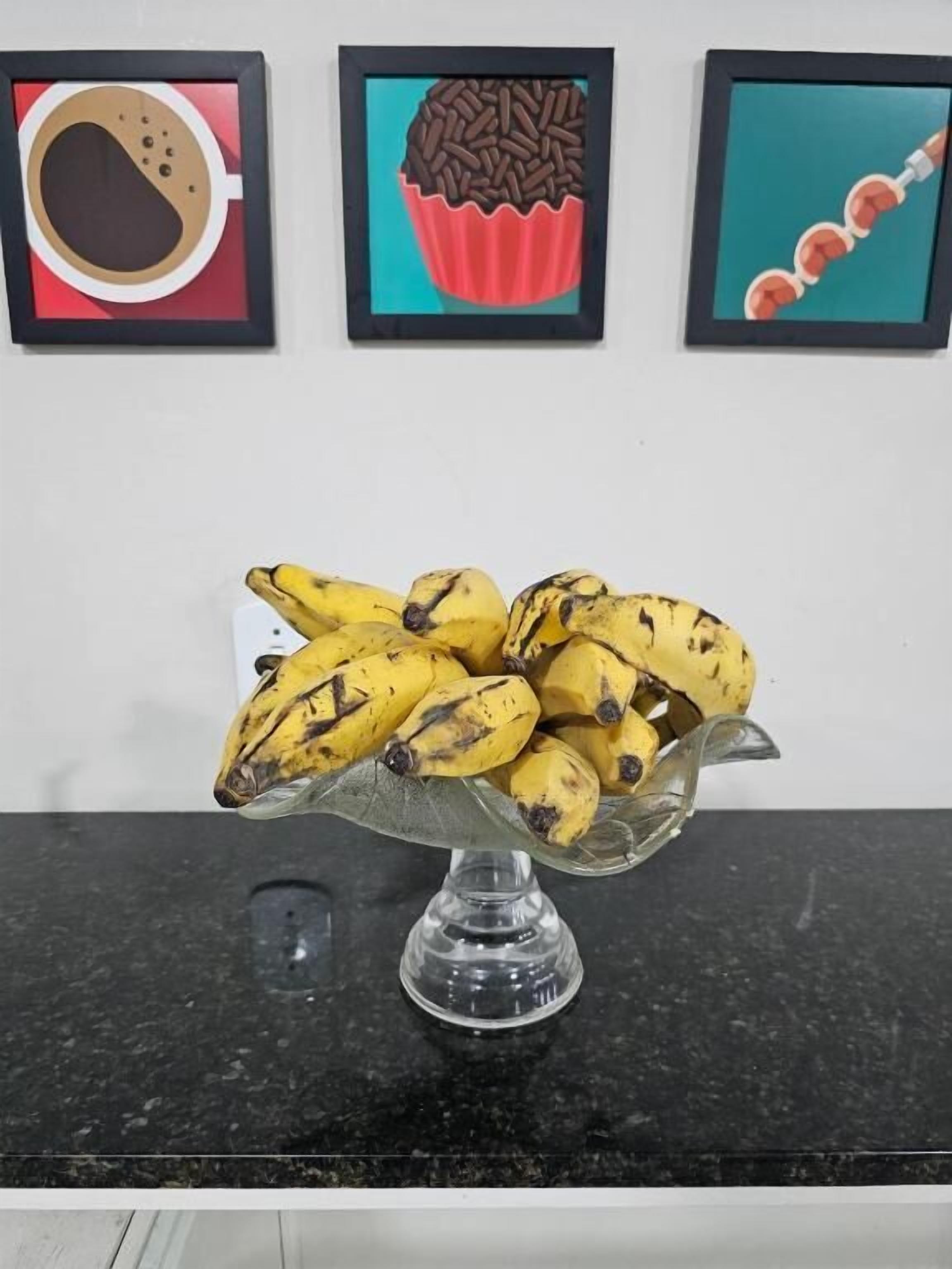 Un saladier en verre rempli de bananes, posé sur un comptoir noir, avec trois tableaux encadrés au-dessus.