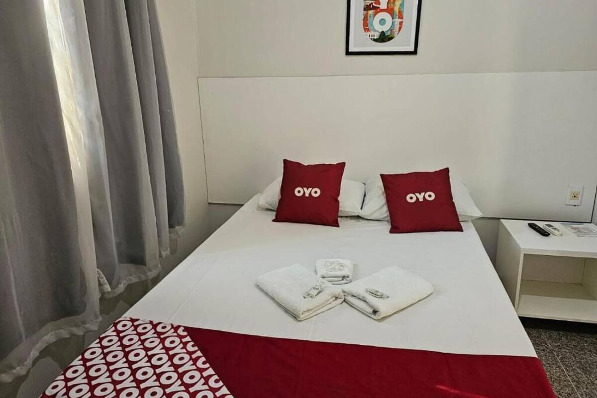 A hotel room with a neatly made bed, two red pillows with the brand name, a framed artwork on the wall, and a bedside table with a remote control.