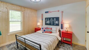 A bedroom with a bed, a window, a red nightstand, a lamp, and a wall with patriotic decorations. - Beautiful Studio Cabin Near Greers Ferry Lake! (Higden)