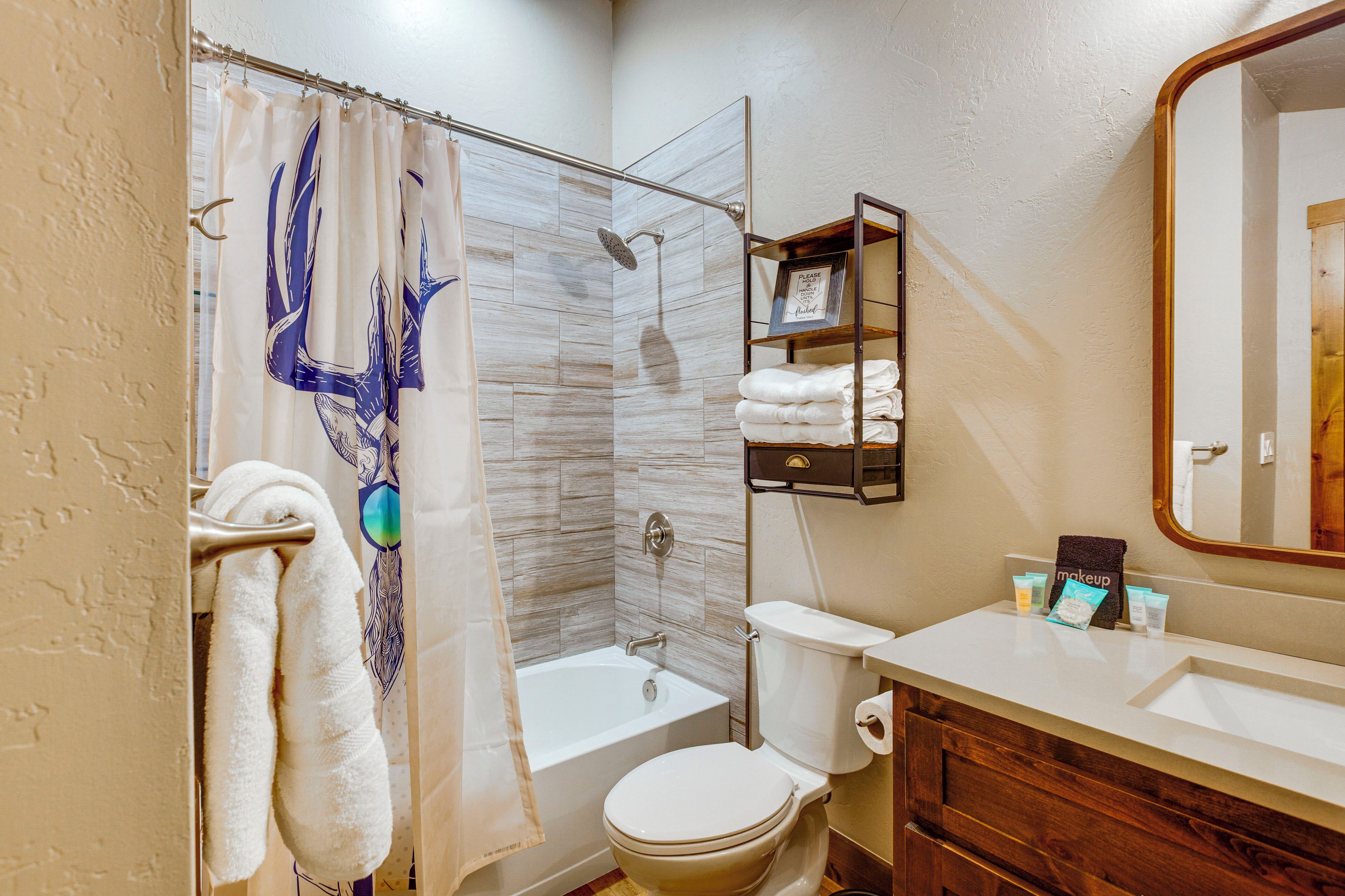 A bathroom with a shower curtain, a bathtub, a toilet, a vanity with a mirror, and a shelf with towels and toiletries.