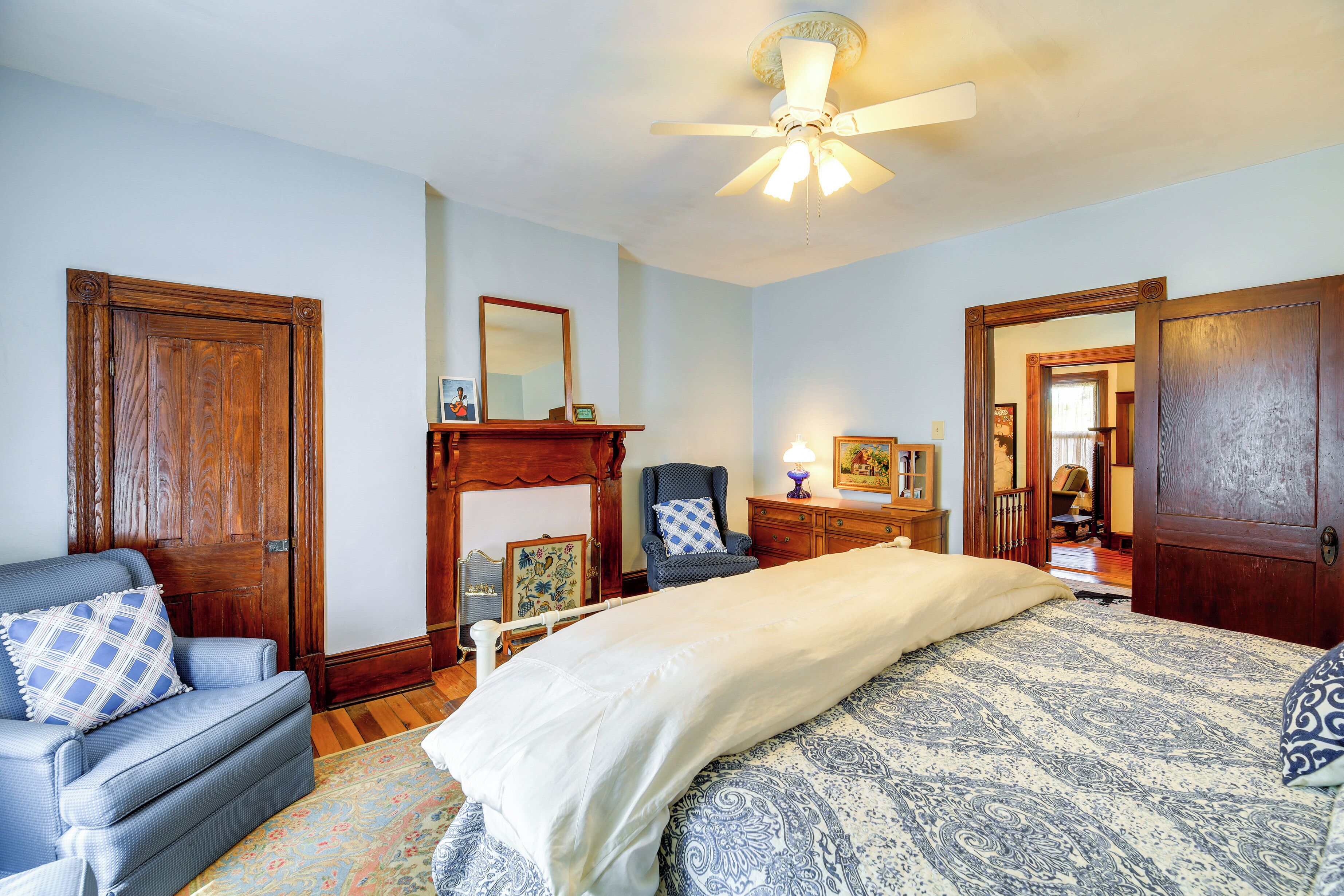 A bedroom with a large bed, a wooden wardrobe, a blue armchair, and a ceiling fan.