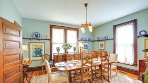 Une salle à manger avec une table en bois dressée pour quatre personnes, entourée de chaises, d’un tapis à motifs floraux et d’un lustre.