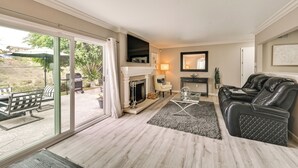 Una sala de estar con chimenea, puerta de vidrio que da a un patio exterior, un sofá de cuero negro y una mesa de centro de vidrio.