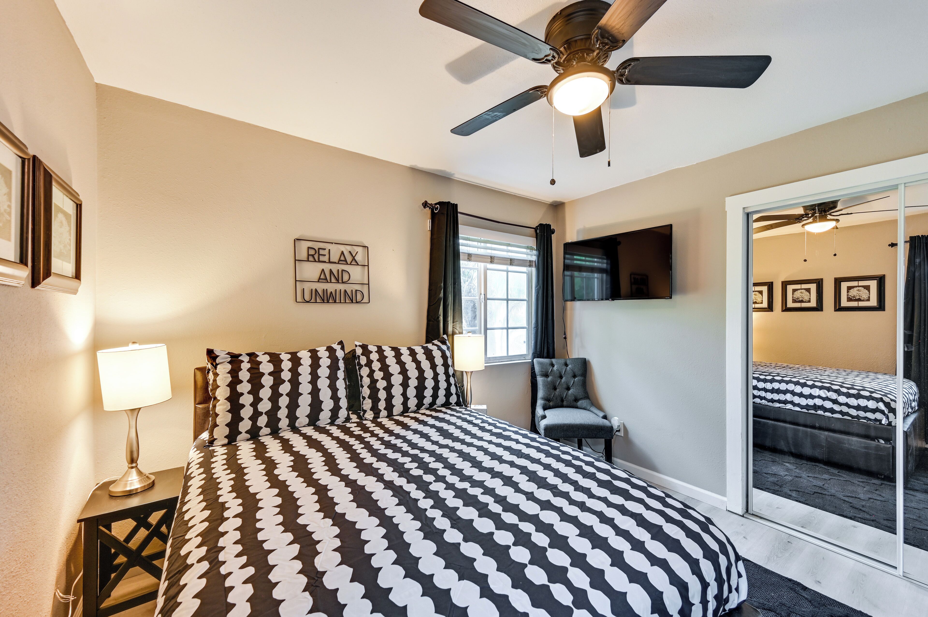 Une chambre à coucher avec un lit, une chaise, un ventilateur de plafond, une fenêtre avec des rideaux et un miroir qui reflète le lit.