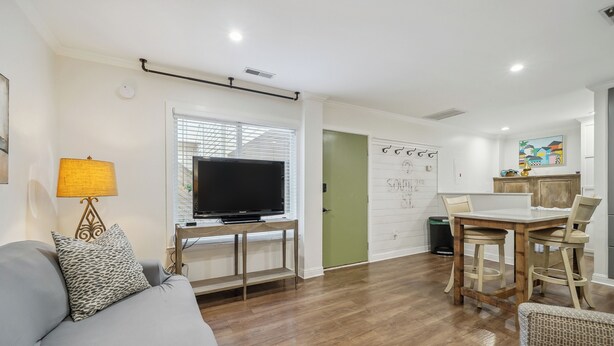 A living room with a sofa, a TV on a stand, a dining table with chairs, and a kitchen area with cabinets and a countertop.