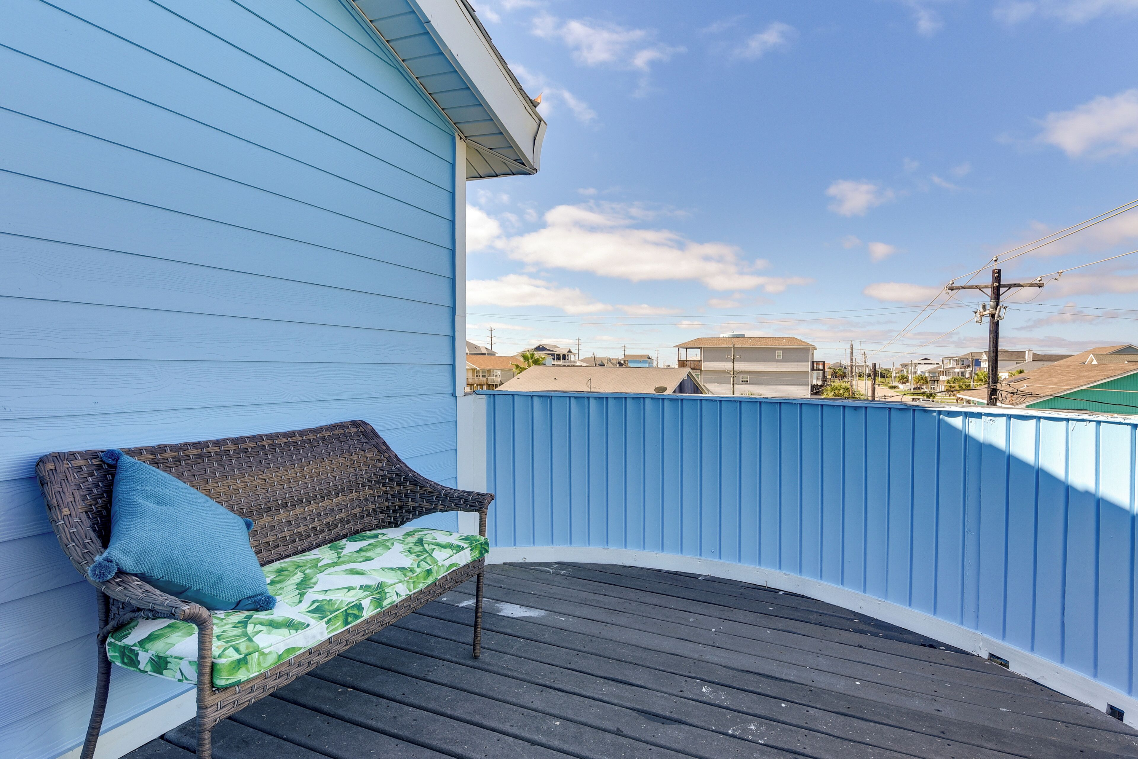 A balcony with a wicker chair and a cushion, overlooking a residential area with houses and a blue fence.