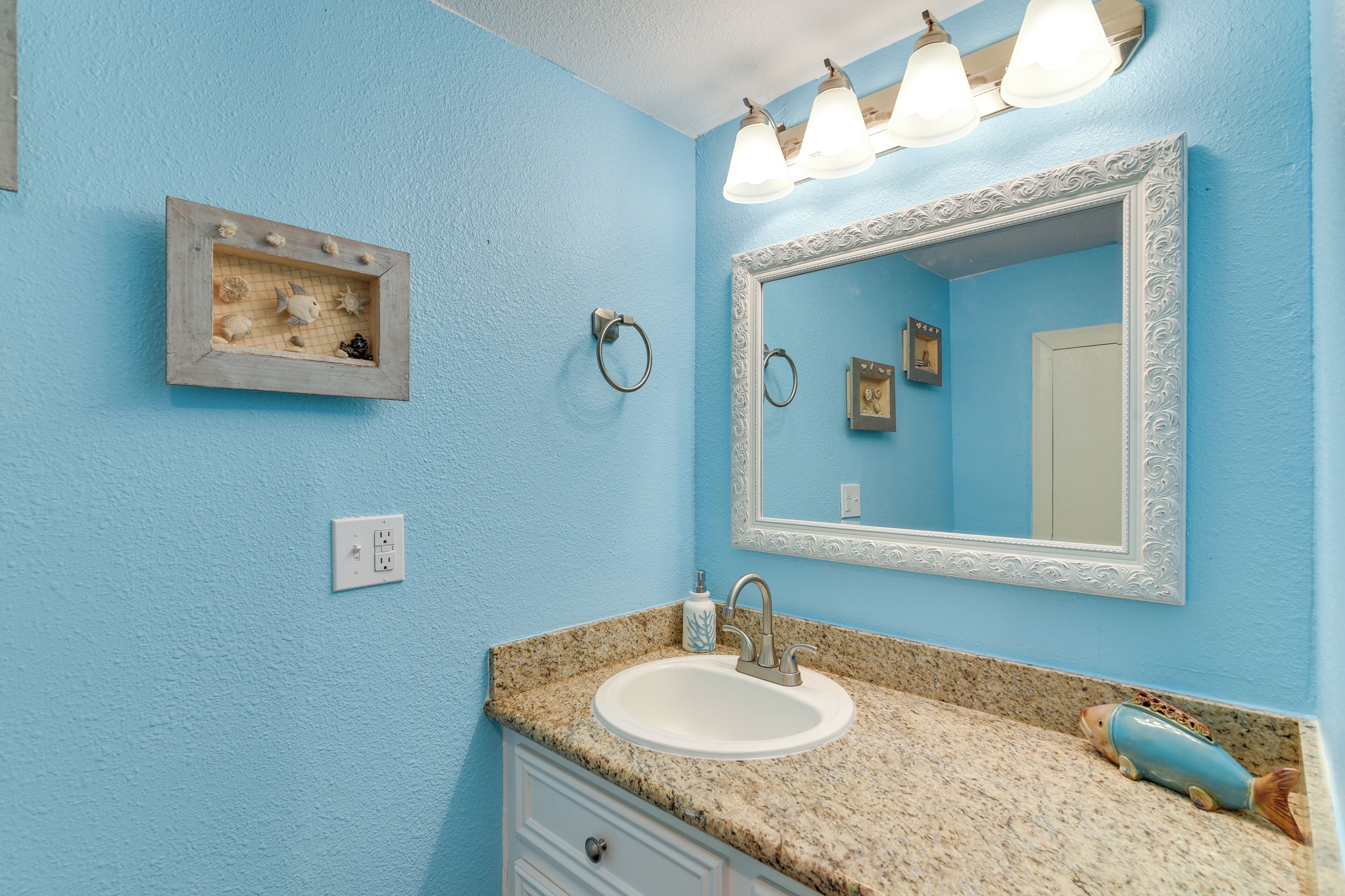 A bathroom with a blue wall, a white sink, a granite countertop, a mirror with an ornate frame, and a small decorative fish on the countertop.