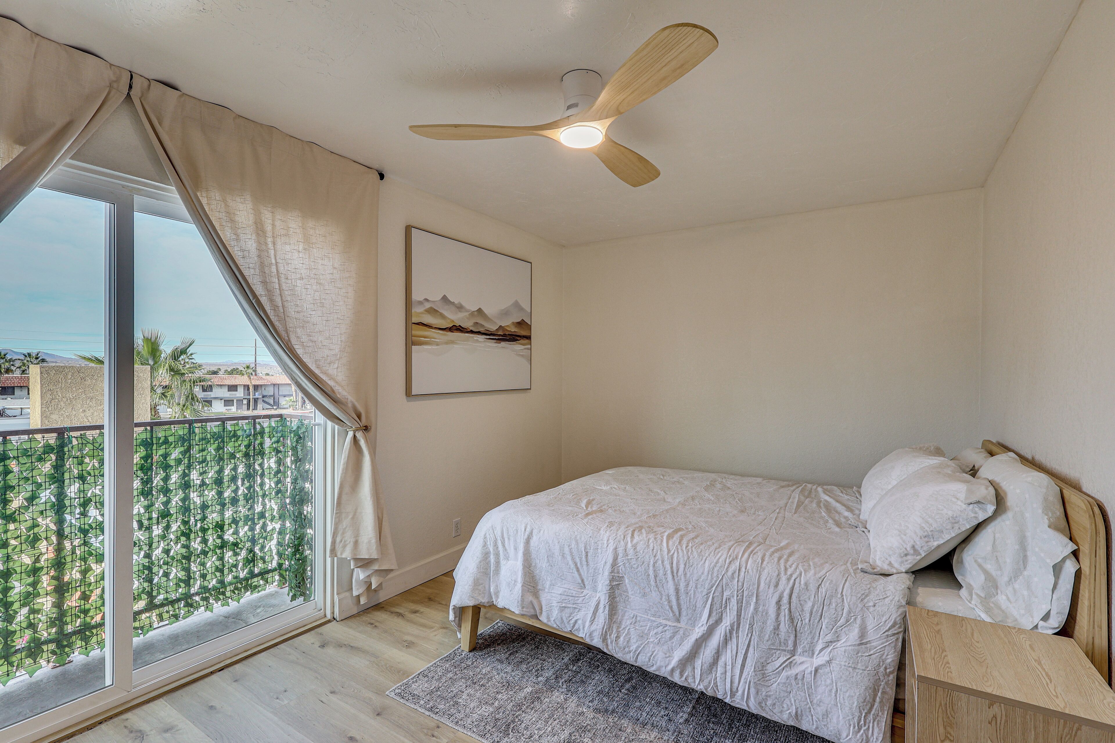 A bedroom with a bed, a ceiling fan, a window with curtains, a framed picture, and a view of the outside.