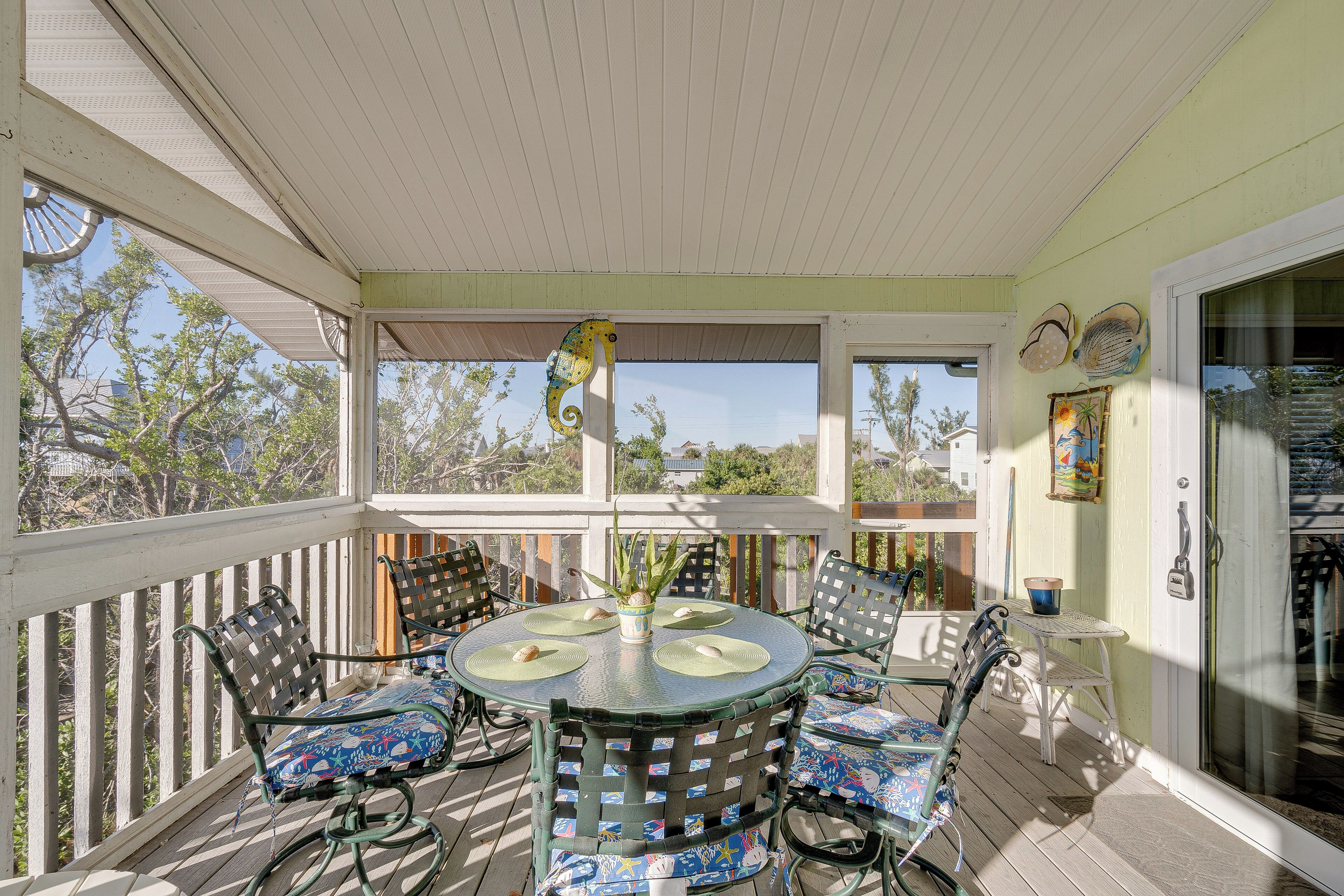 A covered outdoor patio with a round table and chairs, a potted plant, and a view of trees and buildings in the background.