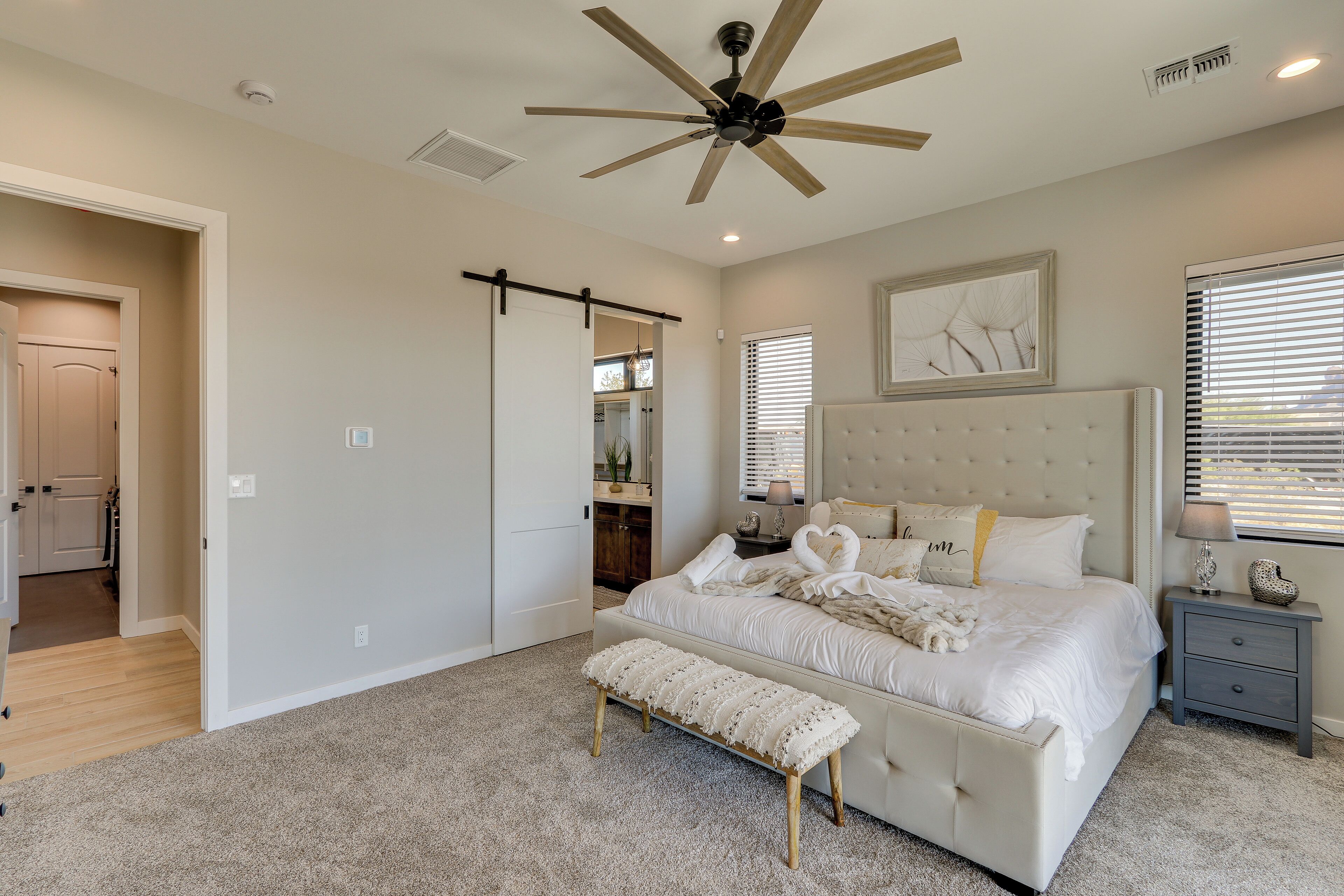A bedroom with a large bed, a ceiling fan, a bench, a nightstand, and a window with blinds.