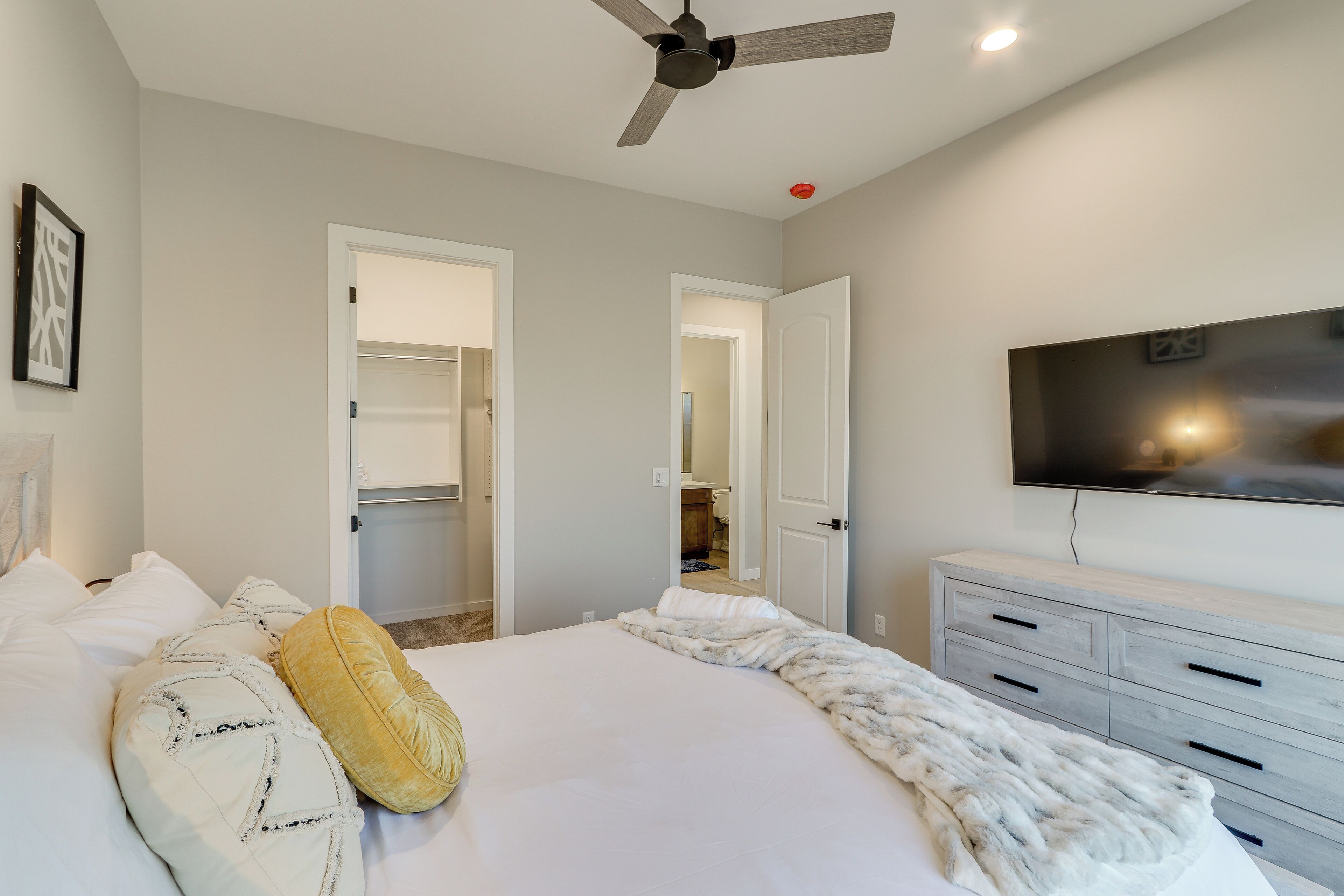 A bedroom with a bed, a ceiling fan, a TV mounted on the wall, and a dresser.