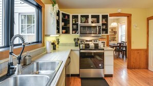 A modern kitchen with a stainless steel sink, built-in microwave, and oven. There is a window with a view of the outside, and a dining area with wooden flooring and chairs. - Charming Bangor Home w/ Deck < 1 Mi to Downtown (Bangor)
