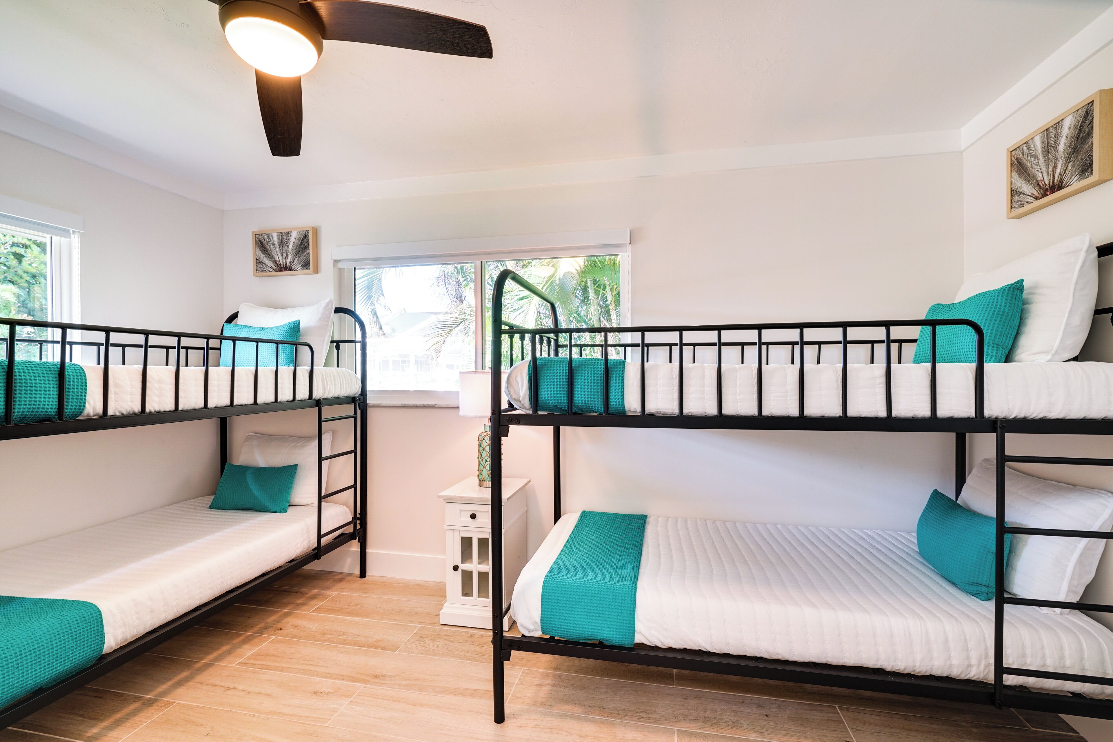 Une chambre avec un lit superposé, dotée de linge de lit blanc et d’éléments de décoration turquoise, d’un ventilateur de plafond et d’une fenêtre donnant sur un paysage verdoyant.