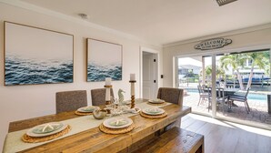 A dining area with a wooden table set for four, surrounded by chairs. There are two framed pictures on the wall, a sign reading "WELCOME TO THE BEACH", and a view of the pool area through glass doors. - Waterfront Fort Myers Home: Private Pool & Dock (Fort Myers)