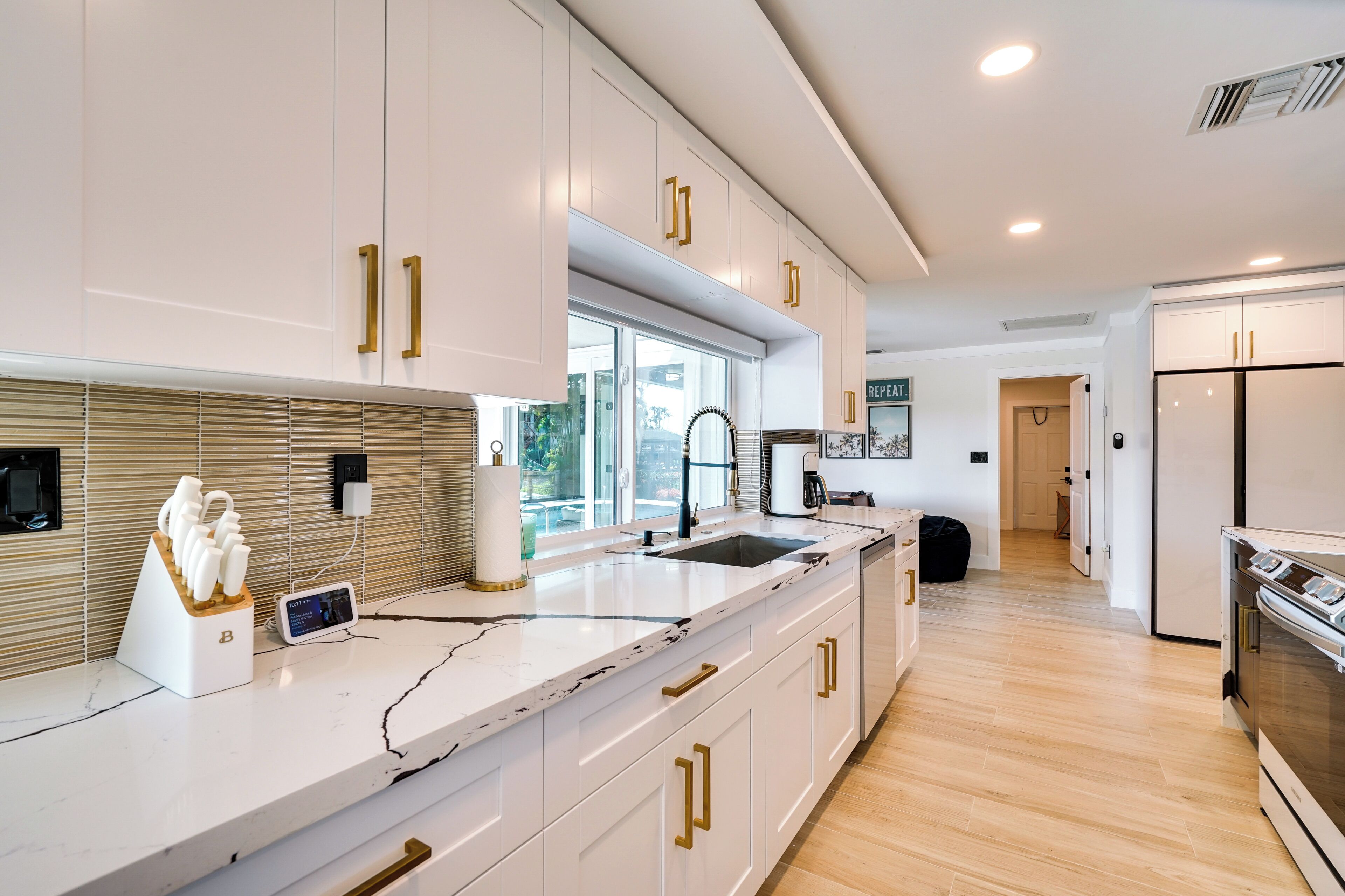 A modern kitchen with white cabinets, gold handles, a marble countertop, and a large window.