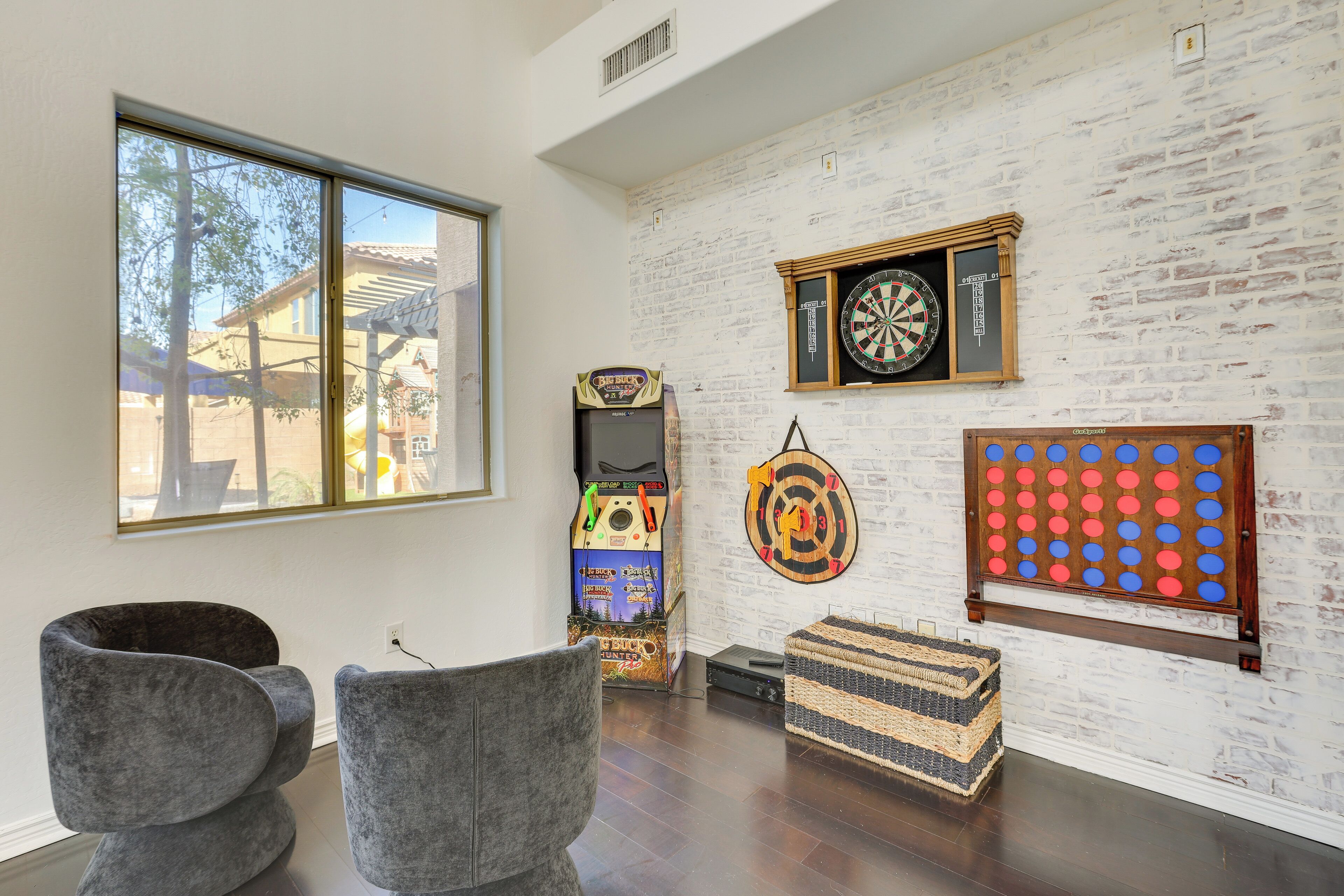 A room with a white brick wall, a dartboard, a pinball machine, and two grey chairs.