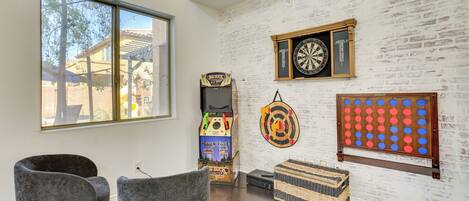 A room with a white brick wall, a dartboard, a pinball machine, and two grey chairs.