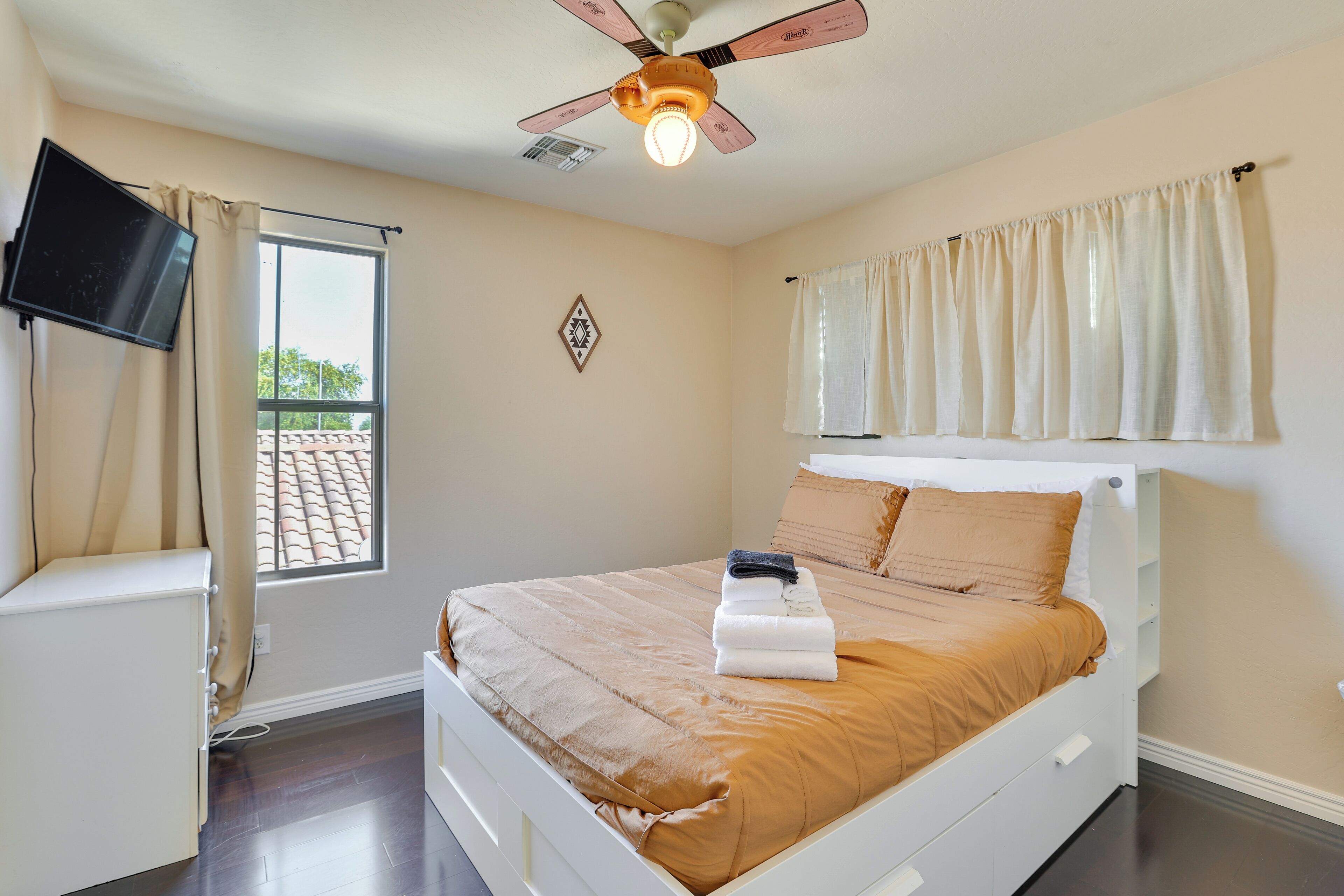 Une chambre à coucher avec un lit, un ventilateur de plafond, une fenêtre avec des rideaux et un téléviseur fixé au mur.